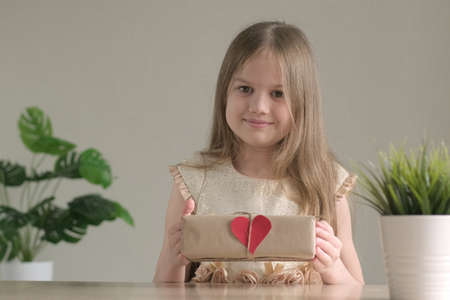 Cute little girl smiling holding handmade stylish simple gift box in craft paper at home with small red heart. Happy child girl with present for Valentines Day. DIY, zero waste concept.の写真素材