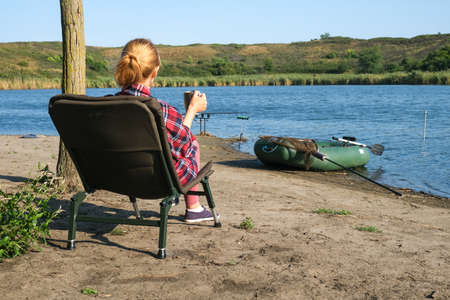 Blonde woman in cozy blanket enjoying hot drink in the morning near the river. Summer vacation, fishing, relax, pleasure outdoorsの写真素材
