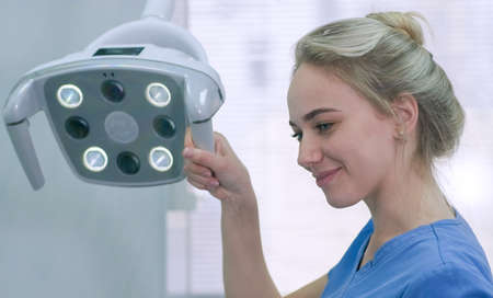 Pretty female nurse adjusting dental light in clinic. Dentist assistant prepares lighting equipment before treatment and smiling at patient. Health care, medicine and dentistry businessの写真素材