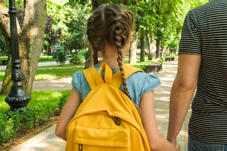 Mother leads the hand of little girl an elementary school student. Mom and daughter walk through the park alley. Woman and child with yellow backpack behind the back, back to schoolの写真素材