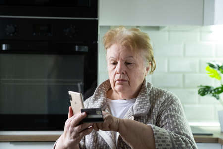 Serious focused elderly woman shopping online with credit card using smart phone at home. Elderly...