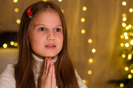 Kid girl is wishing something for Christmas close up. Kid is praying in decorated room on background of Christmas tree and glittering lights. Holidays, childhood, children dreams and family conceptの写真素材