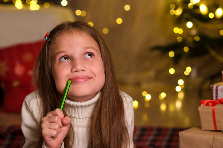 Cute child girl is writing letter to Santa Claus in decorated room for Christmas. Close up kid making wishes near gifts on background of Christmas tree. Holidays and celebrations conceptの写真素材