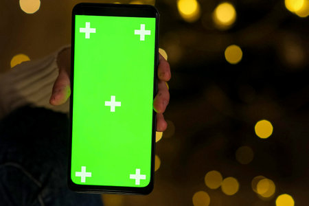 Hand holding Chroma Key Green Screen in vertical Position without touching, swiping at Christmas evening. Using phone with green mock-up, surfing the Internet, watching content videos, appsの写真素材