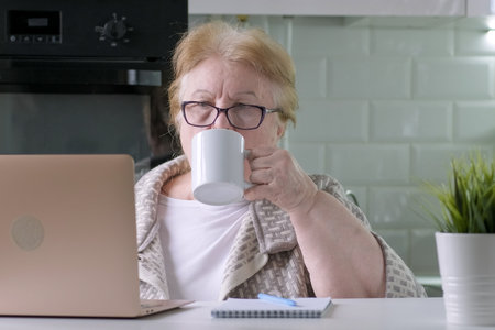 Elderly woman drinking coffee or tea while working on computer laptop at home. grandma freelancer. Checking life insurance or savings financial account, paying bill on bank online app, planning budgetの写真素材