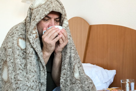 Sick middle aged man sitting on bed at home and treats flu and colds with medicinal drink. Middle...