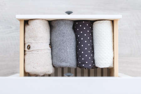 Warm knitted clothing in shades of gray and white is neatly folded in a drawer of a wooden chest of drawers on a gray backgroundの写真素材