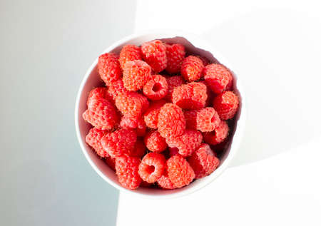 Fresh raspberries in ceramic bowl on white table. Close up photo with sunlight.