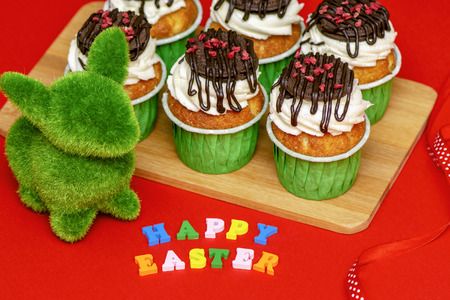 Easter bunny made of grass and delicious bright cupcakes on a wooden board on a red background