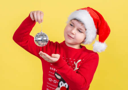 A beautiful 10-year-old boy in a Santa Claus hat holds a Christmas disco ball on a yellow background. Isolate.の写真素材