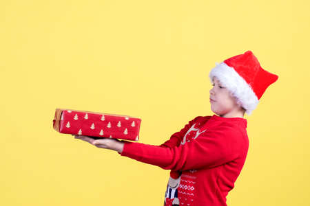 A surprised emotional child in a Santa hat holds a Packed Christmas gift box on a yellow background in the Studio. Copy space.の写真素材