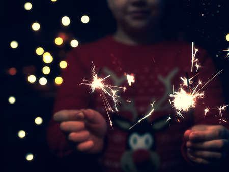 Christmas party. A child holds a lit firework on a shiny background with bright gold Christmas lights. New years eve. Dark concept.の写真素材