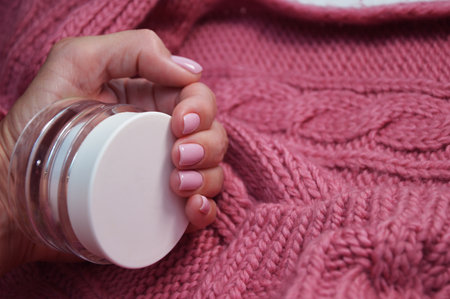 White jar with lotion, moisturizer in a female hand with beautiful nails against the background of a soft and warm bright pink scarf, sweater. Professional manicure. cosmetics. mockupの写真素材
