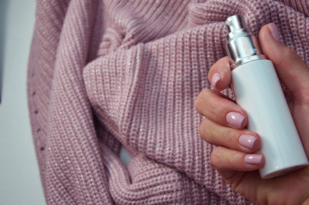 A woman's hand with a beautiful manicure and a white bottle of cream on the background of a pink knitted sweater. winter skincare.の写真素材