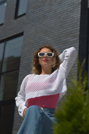 Fashionable confident woman in a knitted white sweater, jeans and trendy sunglasses posing on the...