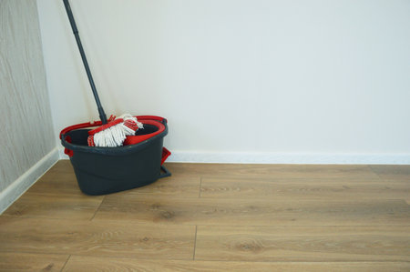 A black bucket with a wringer and a mop stand on a wooden floor near a white wall. Accessories for...