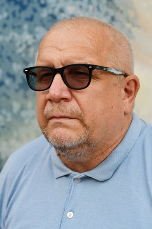 Portrait of an elderly gray-haired man in a blue polo shirt and sunglasses on a light background.