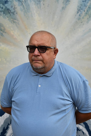 Portrait of an elderly gray-haired man with a beard, wearing sunglasses and a blue T-shirt.