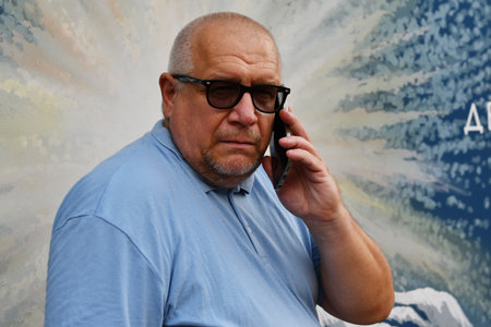 An elderly man wearing glasses and a blue polo shirt and glasses holds a phone to his ear.
