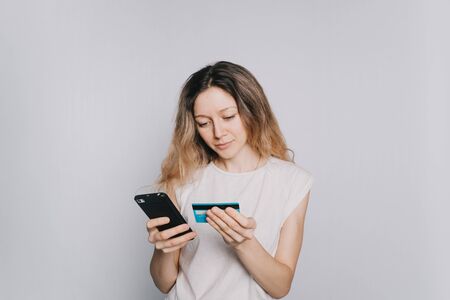 young beautiful Caucasian girl in a white t-shirt makes a purchase using a mobile phone and credit card on white background. Enters a credit card numberの写真素材