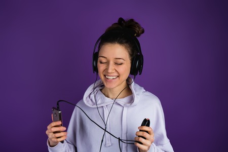 Young beautiful girl with white teeth listening to music on the phone wearing headphones in a sweatshirt on a purple backgroundの写真素材