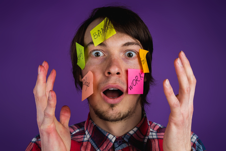 Portrait of a man with stickers on his face shocked by obligations, a man with his hands up in the face in shock from everyday routine, purple backgroundの写真素材