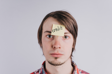 Sad man looks directly, a sticker on his forehead with the word credit. A young guy is upset by debt, credit. Close-up portrait, isolatedの写真素材