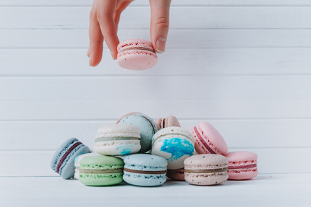 Female hand adds macaroons in a pile of almond cookies on a white wooden background, copy spaceの写真素材