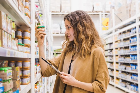 Young beautiful woman with a tablet selects baby food in a supermarket, the girl carefully reads the composition of the productの写真素材