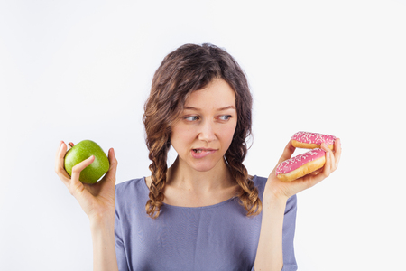 A young woman chooses between a donut and an apple. The concept of harmful nutrition and dietの写真素材
