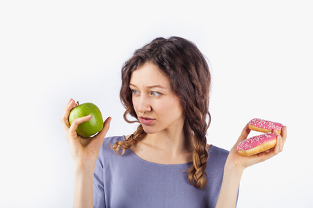 Young woman chooses between a donut and an apple. Concept of harmful nutrition and dietの写真素材