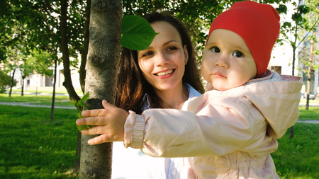Happy mother holds a child in her arms in a city park. Baby is playing with a leaf on the tree at sunset.の写真素材