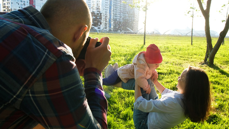 Young father taking pictures of mother and daughter playing in the park at sunset. Happy family photographed in natureの写真素材