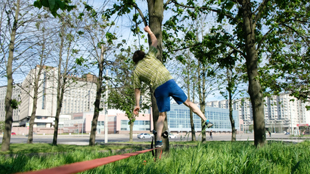 Young European man walks by a tight line in a city park. The guy is balancing on the slackline.