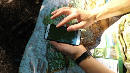 Man tourist sits in the forest and uses an electronic map on the phone, checks the route on a paper...