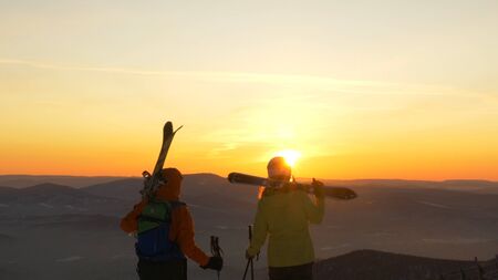 A couple of mountain skiers in love stands on the slope of the mountain and admires the sunsetの写真素材