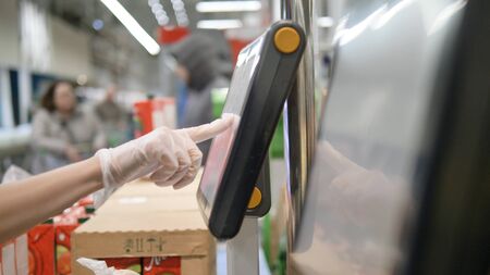 Womans hand in protective gloves weighs vegetables on an electronic scale in a supermarket, selects the desired product on the touch screen. Protective measures against coronavirus.の写真素材