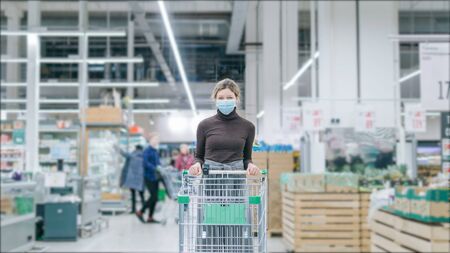 A serious girl in a medical mask with a grocery cart is standing in a supermarket, there are crowds of customers walking around.の写真素材