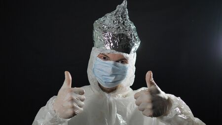 man in a protective suit from coronovirus puts on hat of aluminum foil and throws out sheet with inscription 5G raises his fingers up, on black background andの写真素材