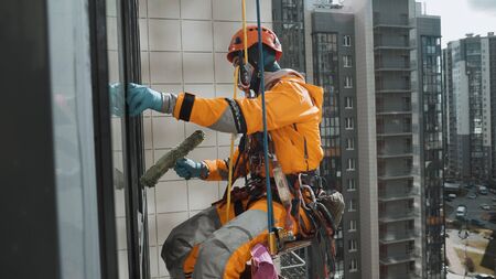 professional industrial climber in helmet washes windows with fur coat at high altitude outside the apartment building in sunny weatherの写真素材