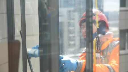 Industrial climber washes windowsin jumpsuit, helmet and gloves in tall megalopolis buildings, close-up slow motion balcony from dust and dirtの写真素材