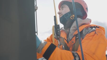 smiling industrial climber in an orange suit and latex gloves washes windows at high altitude...