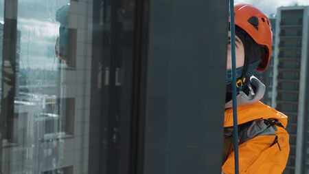 industrial climber window cleaner in orange overalls lathers a glass with a fur coat prepares glass for washing at high altitude, slow motionの写真素材