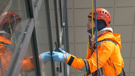 Young industrial climber in orange overalls and helmet screeds windows on a tall building outside hanging on ropes, slow motionの写真素材