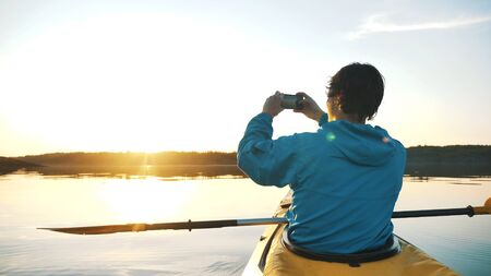 man makes a sunset photo on a smartphone sitting in a kayak on a calm lake, outdoor activitiesの写真素材