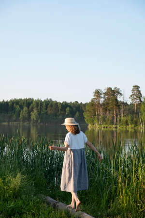 Lovely girl in a straw hat and dress walks barefoot on a log by the lake on the background of tall grass and pine forest on a summer evening..の写真素材