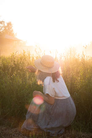 Portrait of a girl from the back in a hat and dress in high grass opposite the suns rays. The girl pushes grass away and looks forward.の写真素材