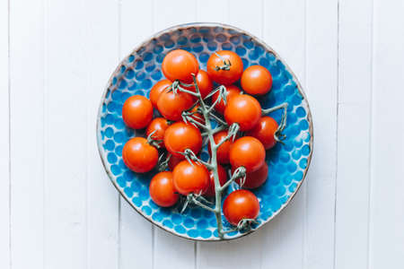 Appetizing ripe cherry tomatoes on a twig in a blue clay plate on a white wooden background. Fresh and healthy vegetarian food.の写真素材