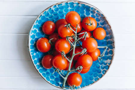 Appetizing ripe cherry tomatoes on a twig in a blue clay plate on a white wooden background. Fresh and healthy vegetarian food.の写真素材
