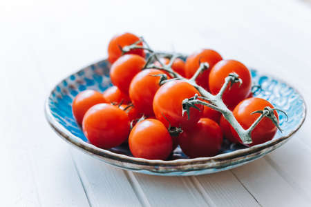 Appetizing ripe cherry tomatoes on a twig in a blue clay plate on a white wooden background. Fresh and healthy vegetarian food.の写真素材